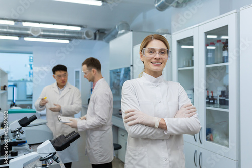 Portrait of a young woman scientist, chemist, doctor standing in the laboratory of the clinic, crossing her arms on her chest and smiling at the camera. In the background, male colleagues.
