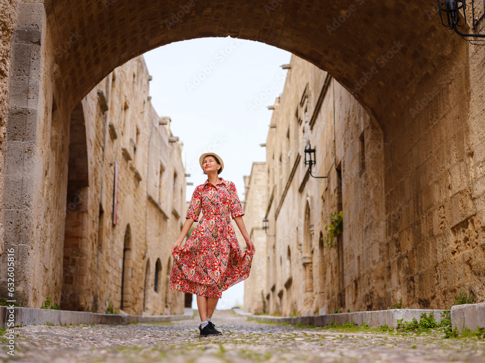 summer trip to Rhodes island, Greece. Young Asian woman in ethnic red dress walks Street of Knights of Fortifications castle. female traveler visiting southern Europe. Unesco world heritage site.