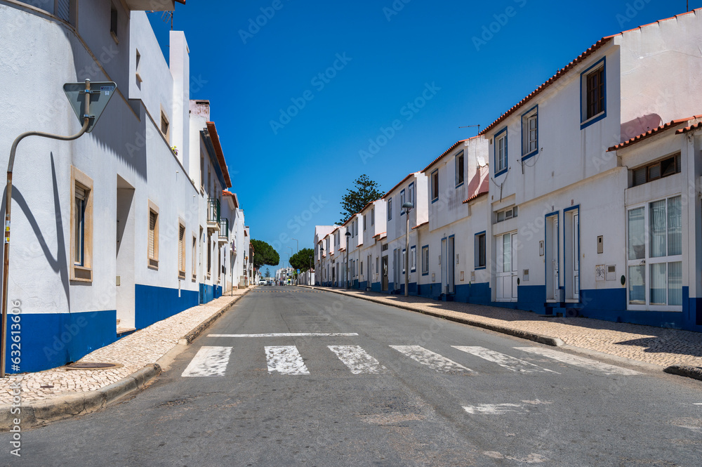 Fototapeta premium Street in Porto Covo, Alentejo, Portugal, with traditional white buildings featuring blue accents on a sunny day.