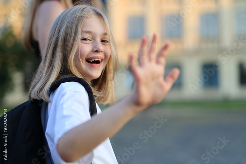 A girl with a backpack waves happily in front of school building on a sunny day.