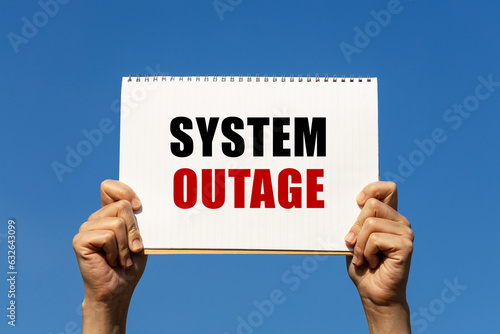 Konstfotografi System outage text on notebook paper held by 2 hands with isolated blue sky background