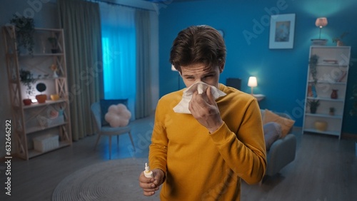 A man with symptoms of a cold stands in the living room close up. A man blows his nose into a paper handkerchief and holds a nasal spray. Home medicine concept.