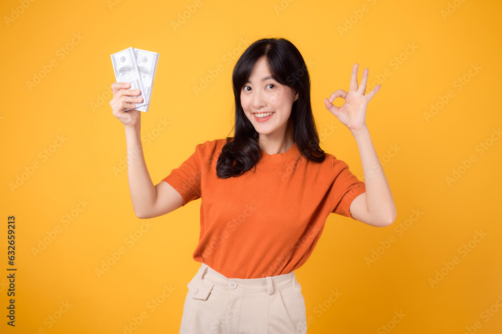 Positive young Asian woman in her 30s, pointing finger to cash money dollars, Okay hand sign, standing on vibrant yellow backdrop.