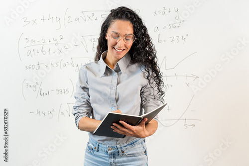 Young Hispanic female school math teacher, college tutor, coach looking at note book, writing and smiling. Background is a white board with math tasks. Concept of education and lesson management.