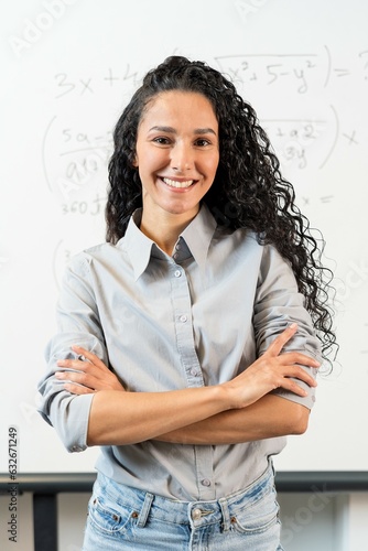 Portrait of a pretty female teacher standing in a classroom with long curly hair of Eastern appearance. Female student in a shirt crossed her arms. Confident look and readiness to start the lesson
