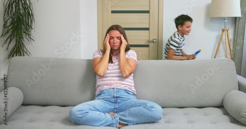 Mother sitting on couch, feeling tired of active children playing running around