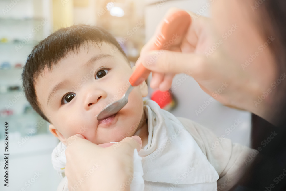Close-up portrait of asian cute baby being fed by her mother using spoon, Mother feeding baby in high chair Close-up portrait of asian cute baby being fed by her mother using spoon, Mother feeding baby in high chair