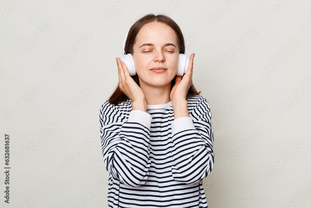 Satisfied woman with brown hair wearing striped casual shirt standing isolated over gray background enjoying favorite tracks with closed eyes keeping hand on headphones. Satisfied woman with brown hair wearing striped casual shirt standing isolated over gray background enjoying favorite tracks with closed eyes keeping hand on headphones.