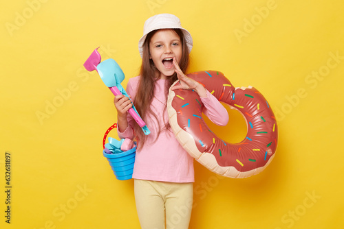 Sunny coast adventure. Excited amazed little cute girl in panama holding beach sandbox toys rake shovel and rubber ring isolated over yellow background screaming making announcement.