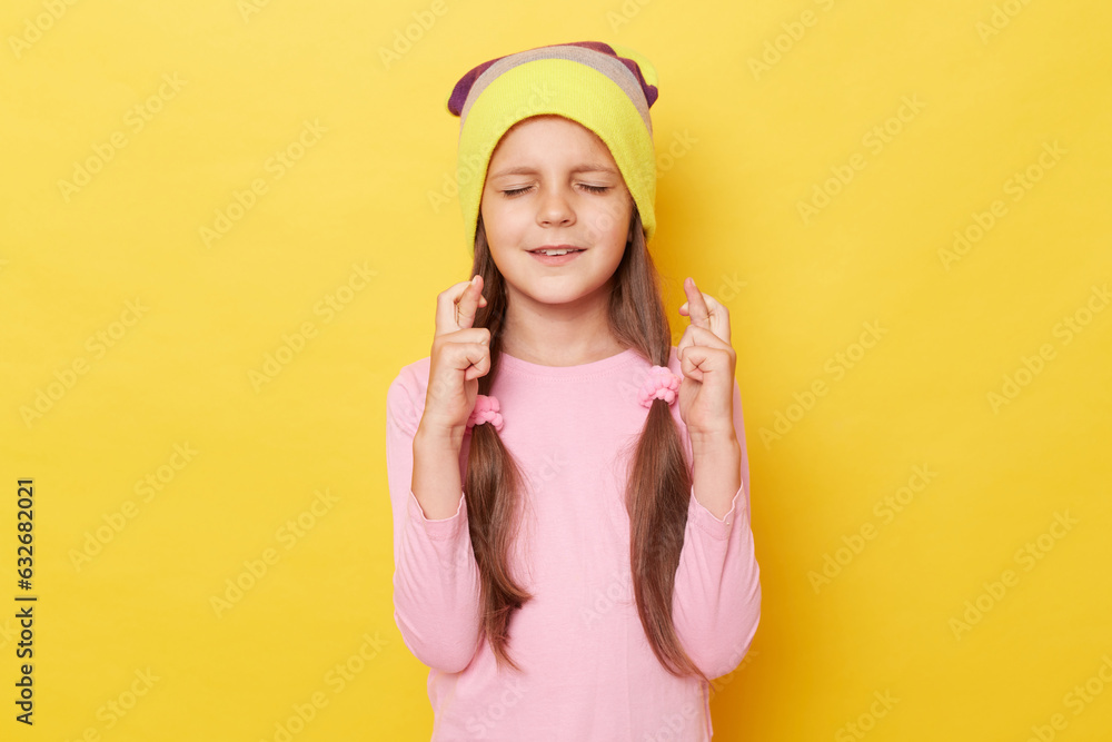 Little girl with ponytails wearing beanie hat standing isolated over yellow background posing with closed eyes crossed fingers making wish being hopeful. Little girl with ponytails wearing beanie hat standing isolated over yellow background posing with closed eyes crossed fingers making wish being hopeful.