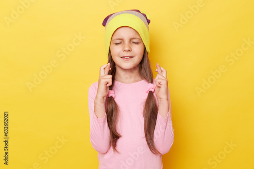 Little girl with ponytails wearing beanie hat standing isolated over yellow background posing with closed eyes crossed fingers making wish being hopeful.