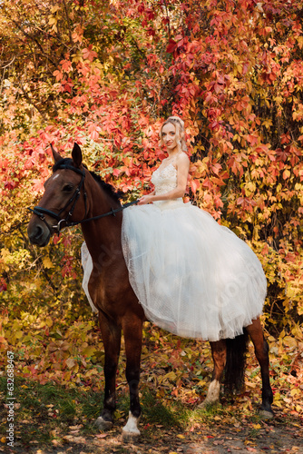 Beautiful bride in white dress stands near a horse in the autumn forest. 