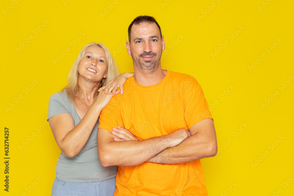 Fototapeta premium Caucasian real couple in their 40s posing happily, the man has his arms crossed and smiles as the woman leans on his shoulder. Isolated on yellow studio background.