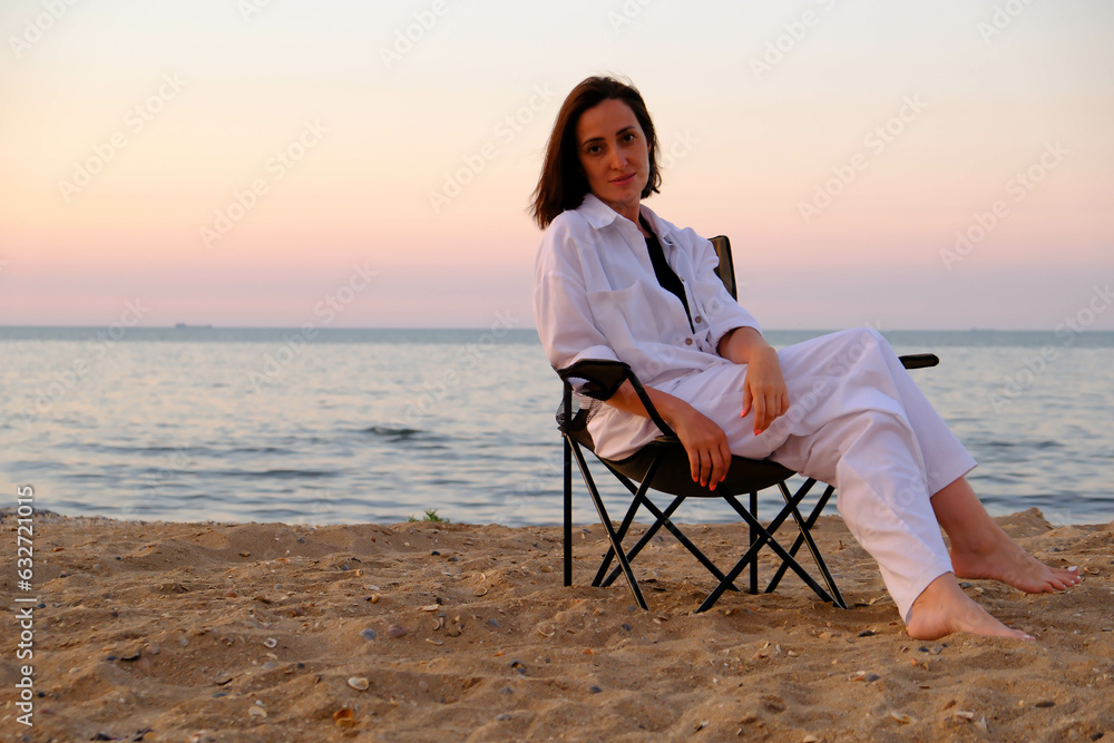 Beautiful cute woman in white suit and straw hat is sitting and relaxing on sea beach coastline.