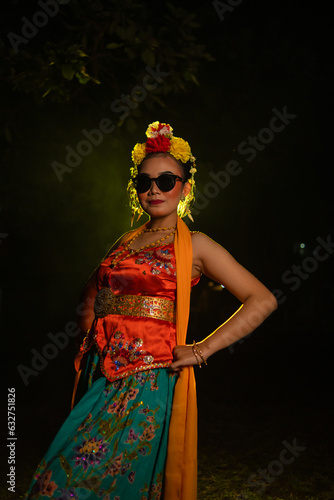 a Sundanese dancer poses in front of a shining light while wearing an orange costume with sunglasses and flowers on her head photo