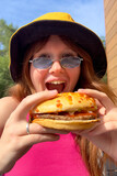 Portrait of young happy woman is eating, biting juicy delicious burger outside cafe looking at camera and smile. Fast junk food, unhealthy eating concept.