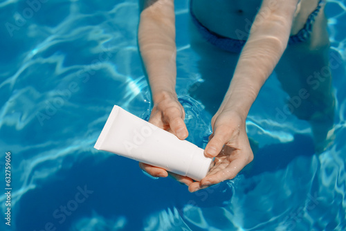 A young woman opens a white tube of make-up day moisturizer in the blue water of a swimming pool. The concept of summer vacation by the water, skin care products. photo