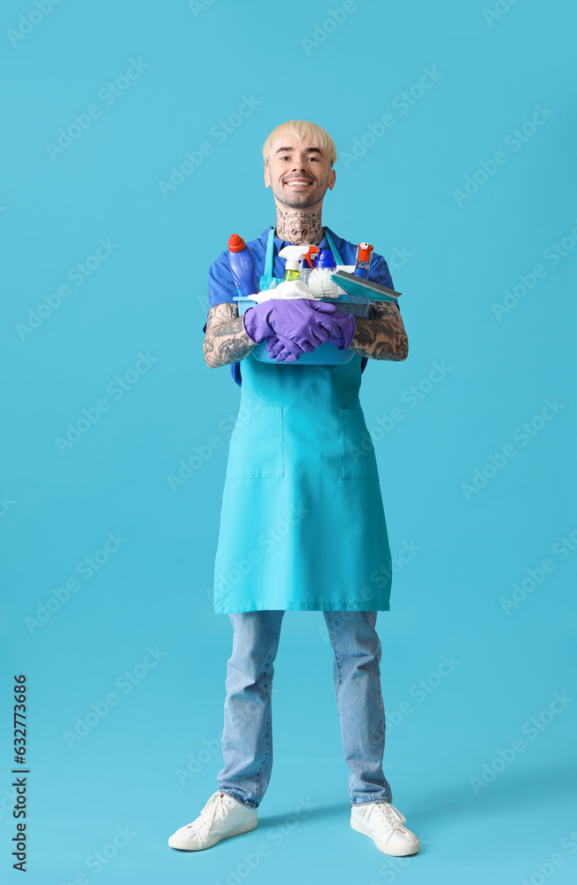 Young tattooed man with bucket of cleaning supplies on blue background