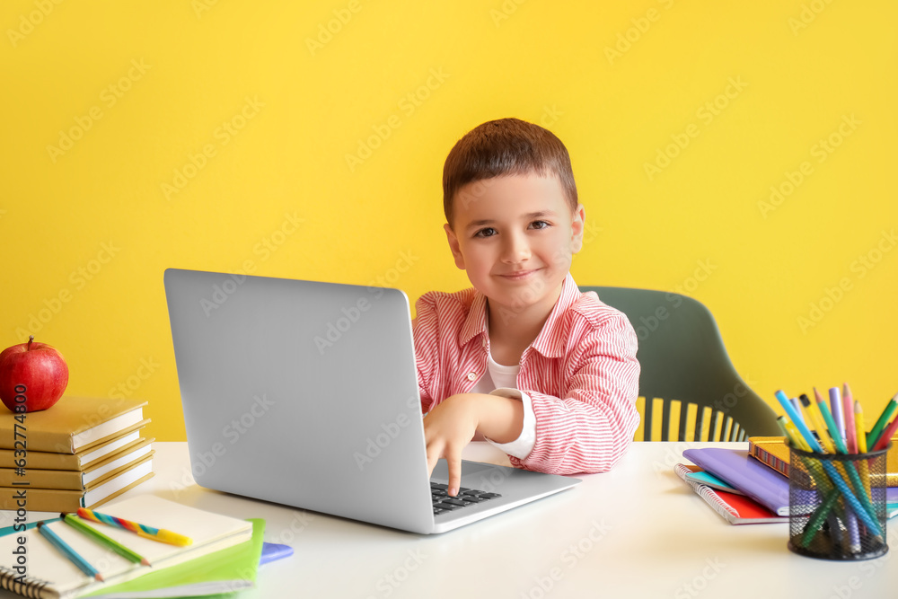 Cute little boy with laptop doing homework at table near yellow wall Cute little boy with laptop doing homework at table near yellow wall