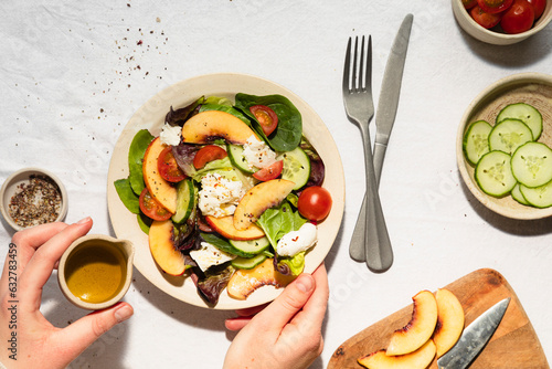 Hands drizzling jug of olive oil over mozzarella and nectarine salad in ceramic bowl, with ingredients surrounding.