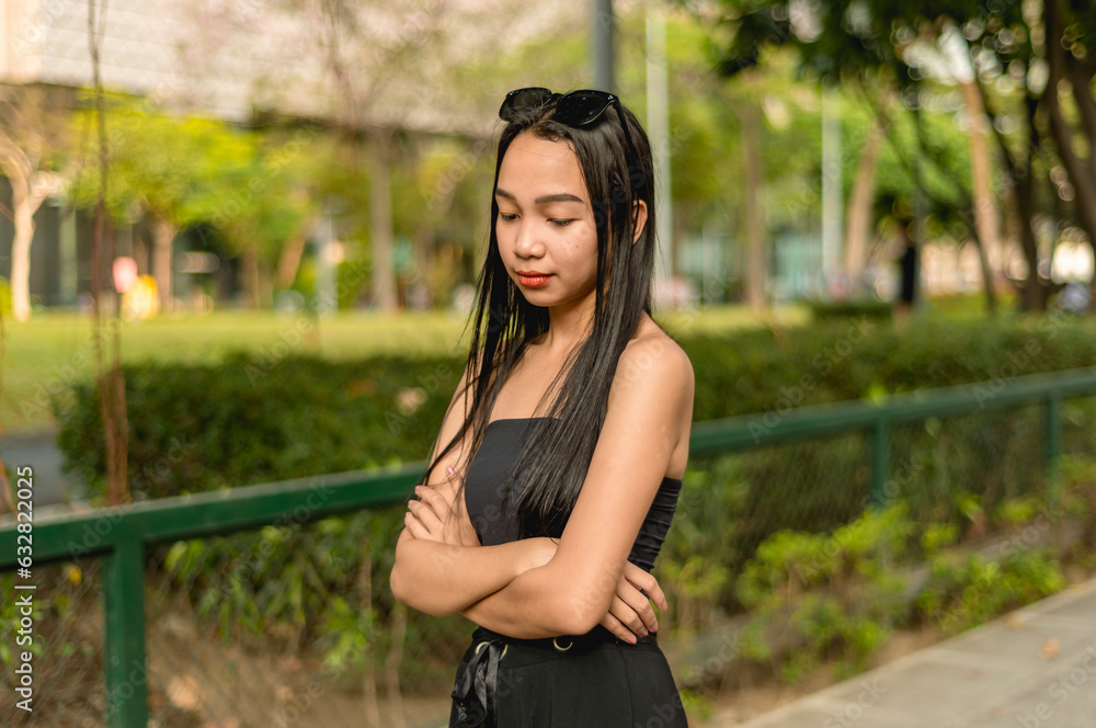 A side view photo of a pretty young lady standing in the park wearing a black strapless tank top and sunglasses, her arms are crossed while looking mad. Trees,bushes and fence in the background. A side view photo of a pretty young lady standing in the park wearing a black strapless tank top and sunglasses, her arms are crossed while looking mad. Trees,bushes and fence in the background.