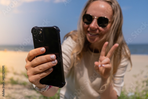 Young woman in sunglasses with shell bracelets holding smartphone. Influencer Using mobile phone for video call selfie waving on the beach seacoast. Holiday vacation advertisement concept. Social