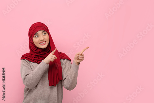 Muslim woman in hijab pointing at something on pink background, space for text