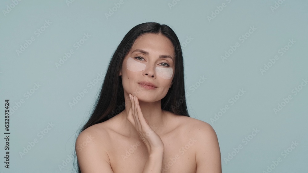 Obraz premium Brunette woman doing facial skin care procedures. Portrait of a seminude woman with hyaluronic patches under her eyes in the studio on a blue background close up.