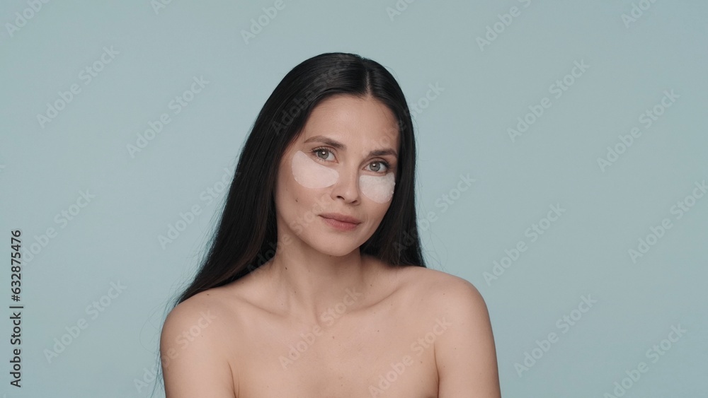 Obraz premium Brunette woman doing facial skin care procedures. Portrait of a seminude woman with hyaluronic patches under her eyes in the studio on a blue background close up.