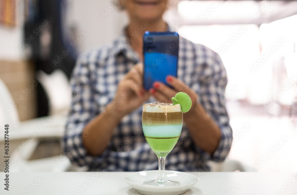 Senior woman using phone sitting at cafe table, a glass of local specialty of Canary island called barraquito in front to her, made with condensed milk, coffee and liqueur Senior woman using phone sitting at cafe table, a glass of local specialty of Canary island called barraquito in front to her, made with condensed milk, coffee and liqueur