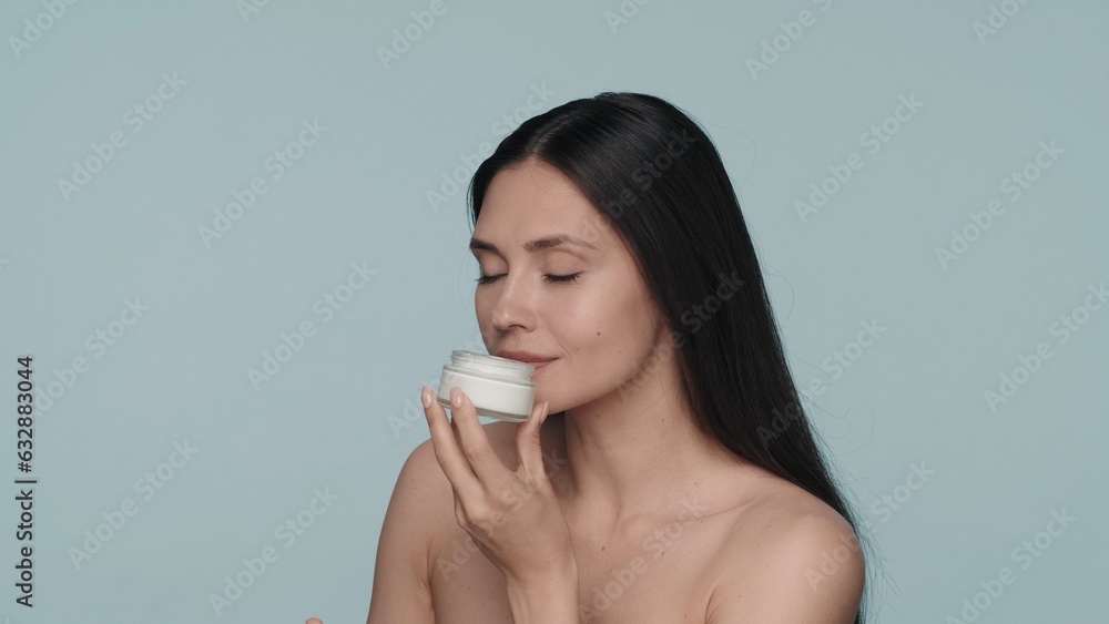 Cream for skin care. A seminude woman holds a jar of cream and inhales its aroma. Woman with cream in her hands in the studio on a blue background close up. Cream for skin care. A seminude woman holds a jar of cream and inhales its aroma. Woman with cream in her hands in the studio on a blue background close up.