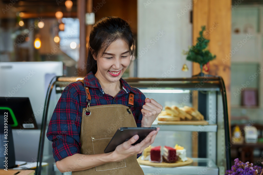 beautiful asian girl Bakery shop owner in overalls Standing smiling and happy when taking orders from customers who order through the app from the tablet. It's the first order from the shop. beautiful asian girl Bakery shop owner in overalls Standing smiling and happy when taking orders from customers who order through the app from the tablet. It's the first order from the shop.