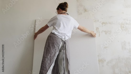 The female artist carries a large blank canvas in her hands and places it against the wall, preparing her work space. Workshop of a contemporary artist.