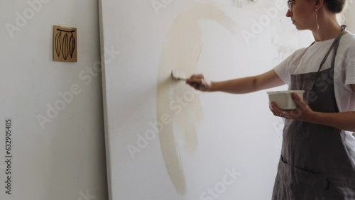 An artist applies beige acrylic paint to a canvas with a brush. A young woman artist works in her workshop. Abstract painting, contemporary art.