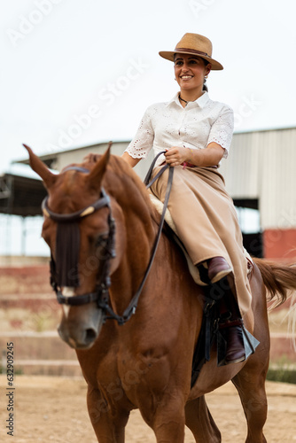 Vertical half-length photo of a girl in elegant clothes on a brown horse with an amazon-type mount on the outside of the stable. The animal is of the Algo-Arabian breed. Cute women riding horses.