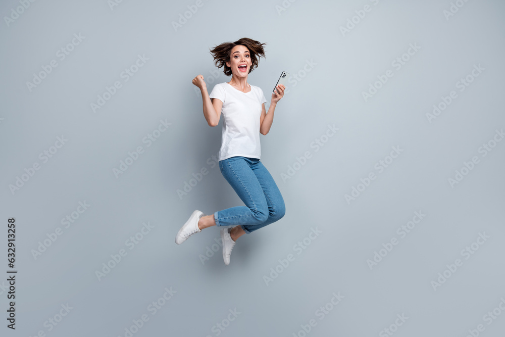 Full size photo of ecstatic overjoyed girl dressed white t-shirt jeans jump hold smartphone win bet isolated on gray color background Full size photo of ecstatic overjoyed girl dressed white t-shirt jeans jump hold smartphone win bet isolated on gray color background