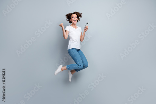 Full size photo of ecstatic overjoyed girl dressed white t-shirt jeans jump hold smartphone win bet isolated on gray color background