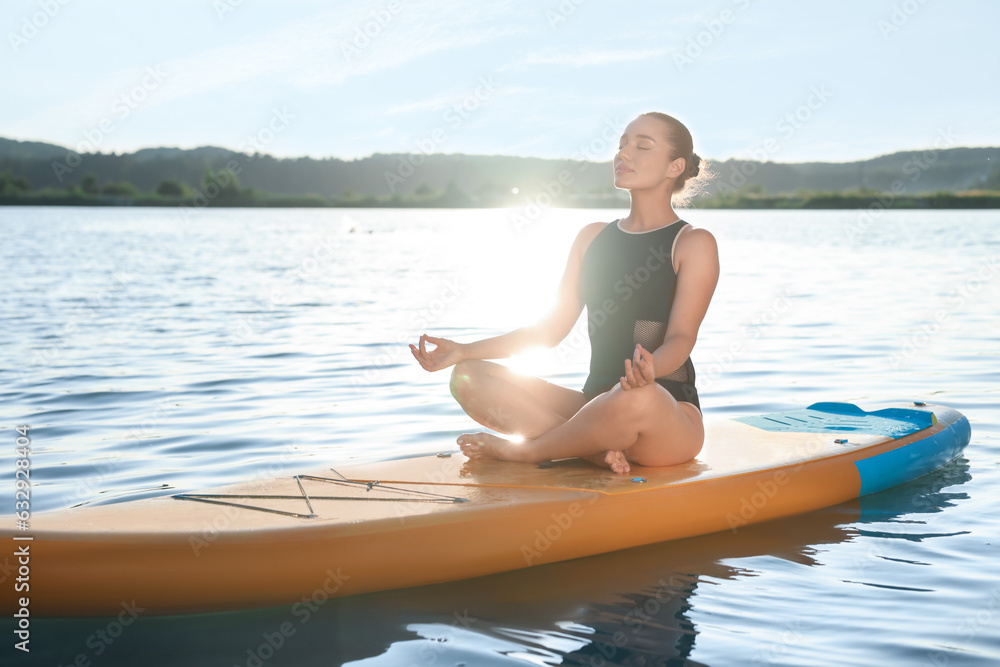 Young meditating yoga on color SUP board on river at sunset