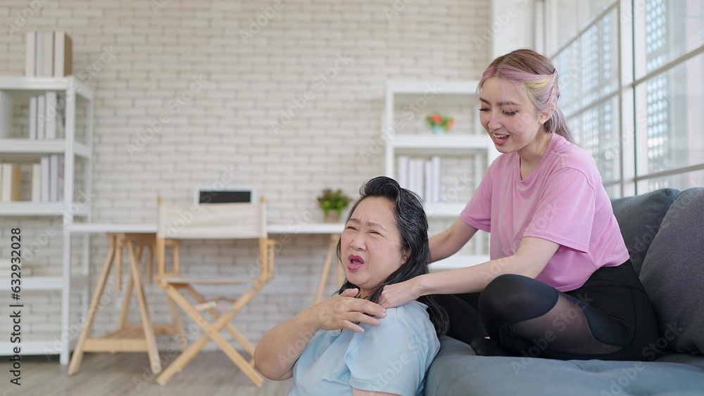 Asian daughter giving shoulder massage to elderly mother while relexing in living room at home. Young daughter giving an elderly mother relaxing massage. Family lifestyle Asian daughter giving shoulder massage to elderly mother while relexing in living room at home. Young daughter giving an elderly mother relaxing massage. Family lifestyle