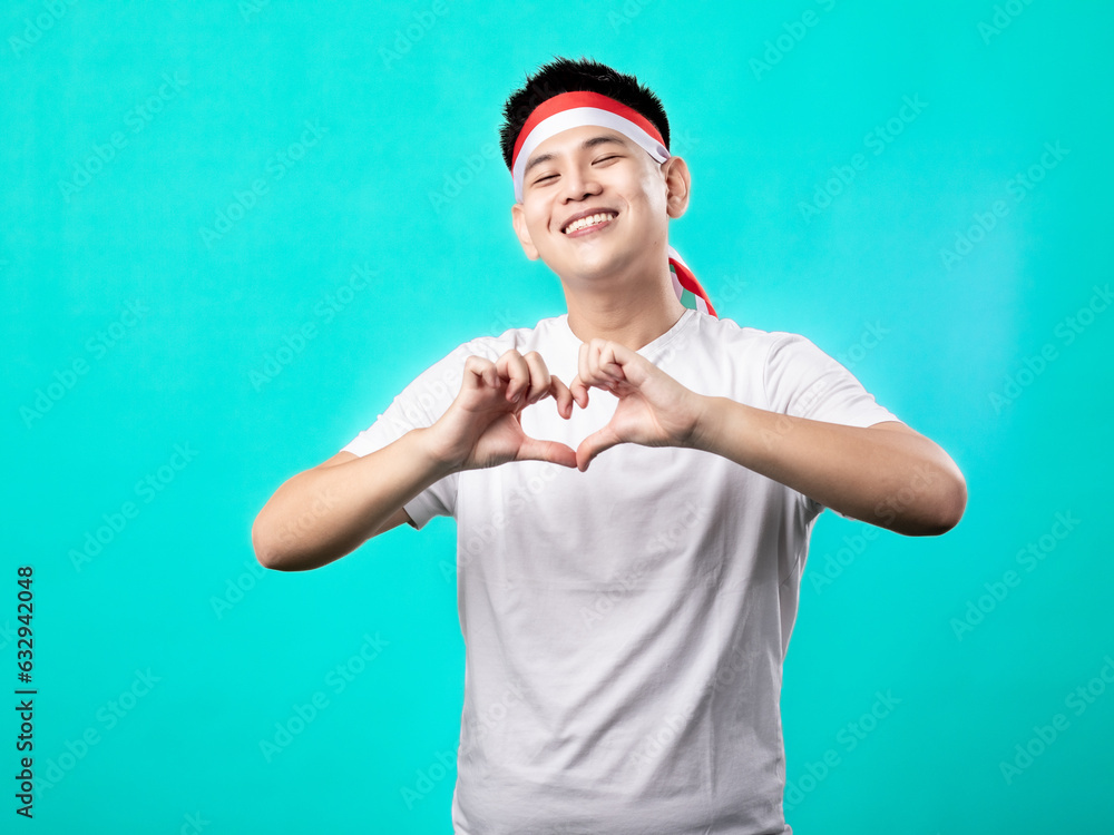 Naklejka premium A portrait of an Asian man celebrating Independence Day, wearing a white shirt with a red and white headband, making a heart-shaped pose in front of his chest, isolated with a turquoise background.