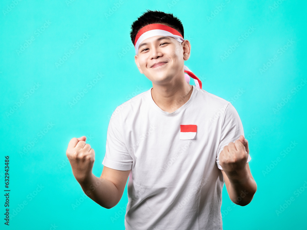 Naklejka premium A portrait of an Asian man celebrating Independence Day, wearing a white t-shirt and a red and white headband, showing a victory pose with his hands raised up. Isolated with a turquoise background.