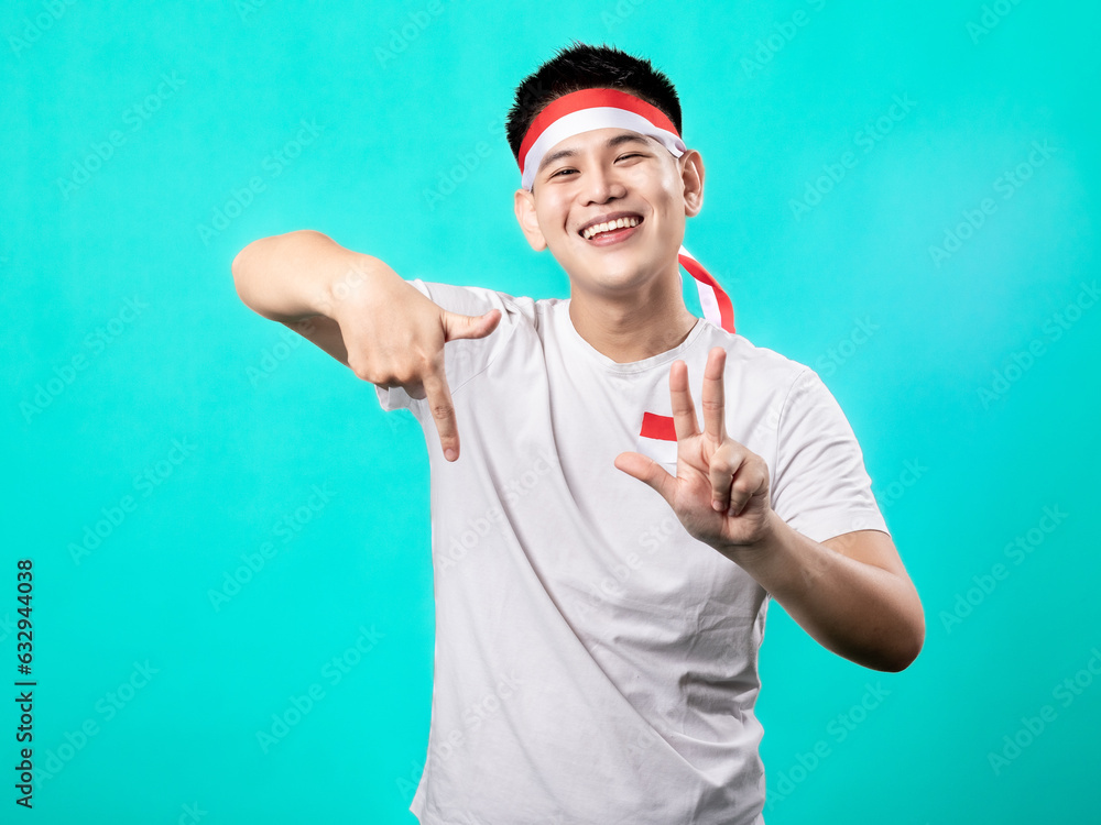 Naklejka premium A portrait of an Asian man celebrating Independence Day, wearing a white shirt with a red and white headband, posing with his fingers forming the number 78, isolated with a turquoise background.