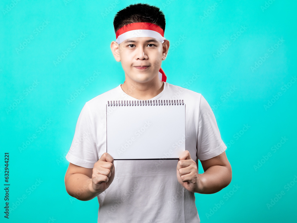 Portrait of an Indonesian Asian man celebrating Independence Day, wearing a white shirt and a red-and-white headband, seen in a pose with an empty paper, isolated with a turquoise background. Portrait of an Indonesian Asian man celebrating Independence Day, wearing a white shirt and a red-and-white headband, seen in a pose with an empty paper, isolated with a turquoise background.