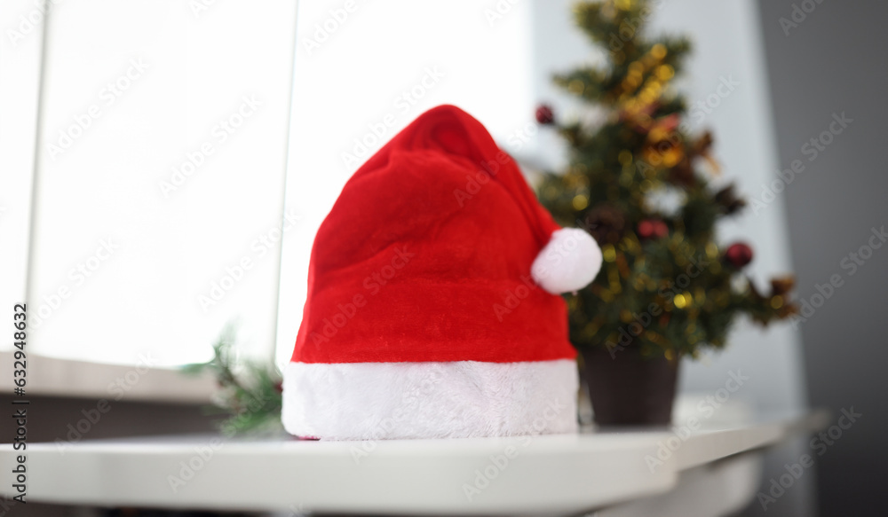 Fototapeta premium Santa claus hat standing near christmas tree closeup. Christmas holidays concept