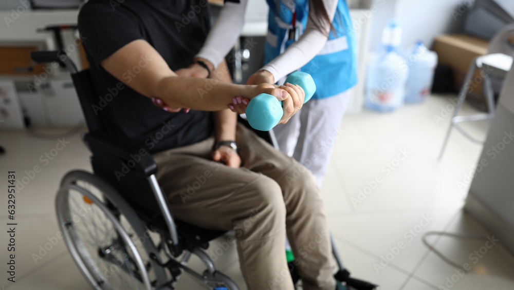 Paralyzed man sit in wheelchair and hold blue dumbbell on his outstretched arm. Femail doctor help patient to knead his hand in clinic. Paralyzed man sit in wheelchair and hold blue dumbbell on his outstretched arm. Femail doctor help patient to knead his hand in clinic.