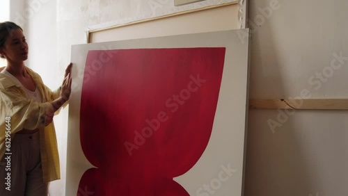 A young artist arranges her paintings in her studio. The woman carries a large canvas and places it against the wall. Workshop of a contemporary artist, abstract painting, art.