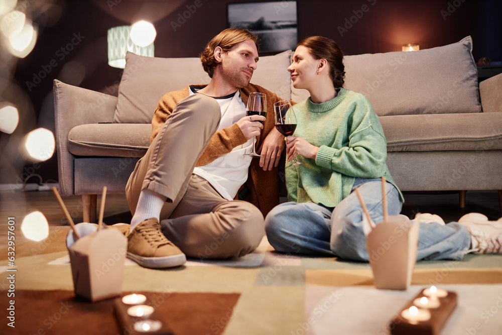 Portrait of young couple enjoying date night at home and drinking wine sitting on floor together, copy space