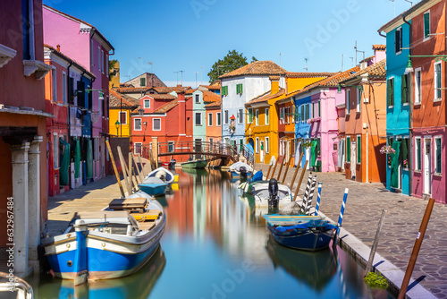 Colourful houses and buildings with reflection in blurred long exposure water of the river. Dock with moored boats on glassy water. Mirror of colors and bright vivid blue sky. Burano, Venice, Italy