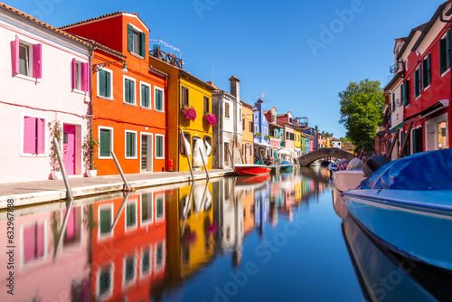 Colourful houses and buildings with reflection in blurred long exposure water of the river. Dock with moored boats on glassy water. Mirror of colors and bright vivid blue sky. Burano, Venice, Italy © Thomas