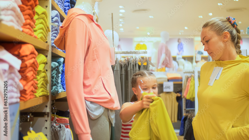 Young mother and child chooses clothes in sports clothing boutique. Cheerful child and mother choosing sports clothes in retail store. Mother and daughter choose looking for clothes in shopping mall Young mother and child chooses clothes in sports clothing boutique. Cheerful child and mother choosing sports clothes in retail store. Mother and daughter choose looking for clothes in shopping mall